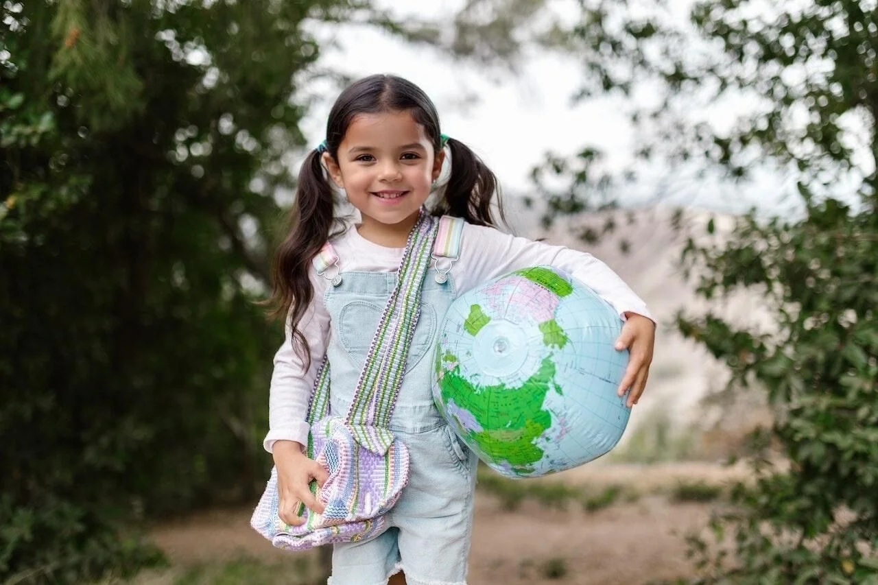 A young girl with pigtails, smiling and holding a large inflatable globe, stands outdoors with trees in the background. The face and hair were AI-generated by EraseID.