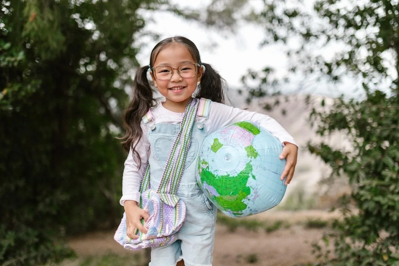 A young girl with glasses, smiling and holding a large inflatable globe, stands outdoors with trees in the background. The face and hair were AI-generated by EraseID.