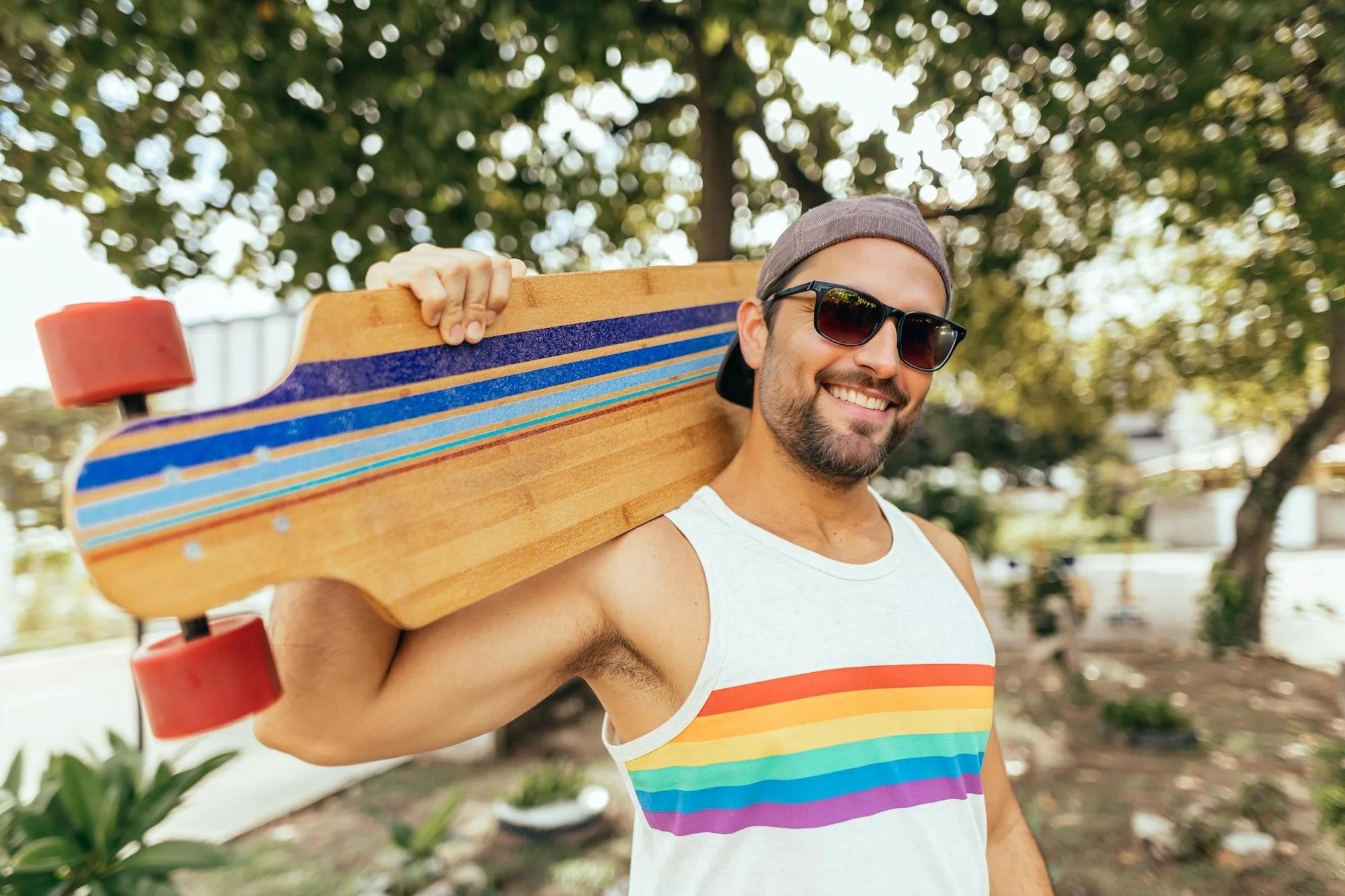 A cheerful man wearing sunglasses and a beanie hat carries a longboard over his shoulder in a park. The face including sunglasses was AI-generated by EraseID.