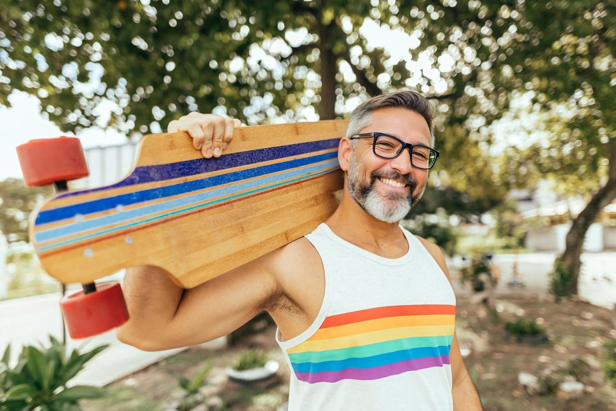 A cheerful man with white beard and hair and wearing glasses carries a longboard over his shoulder in a park. The face including beard, hair and glasses was AI-generated by EraseID.